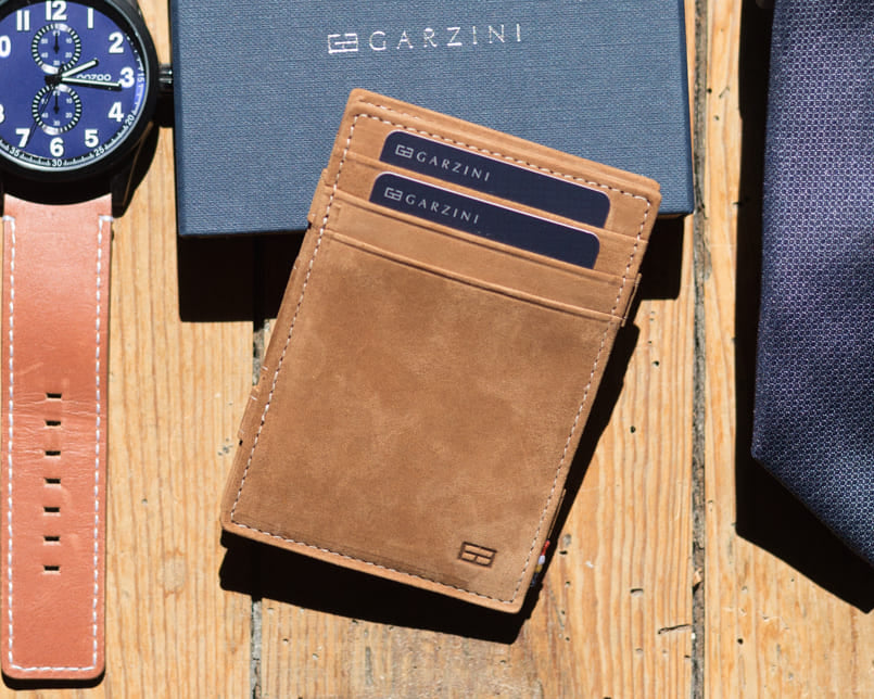 Front view of the Essenziale Magic Wallet Vintage in Camel Brown with three front card slots and two cards displaying the Garzini brand name. The wallet is placed on a Garzini packaging box, with a watch on the left and a tie on the right, both positioned on a wooden table