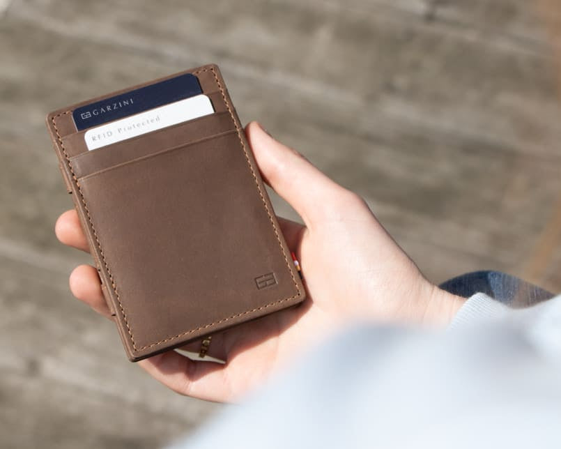 Essenziale Magic Wallet Vintage in Java Brown partially placed inside a bag, alongside a pen, AirPods, and a phone in the background.