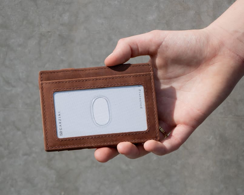 A hand showcasing the Brushed Brown Essenziale Magic Wallet with an ID Window. The hand holds a card inserted into the ID window of the wallet.