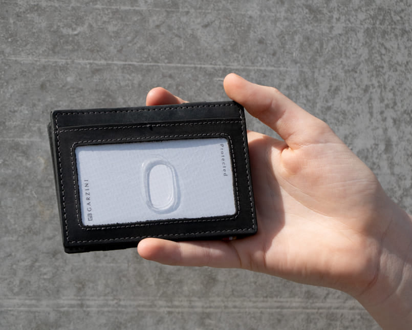 A hand showcasing the Carbon Black Essenziale Magic Wallet with an ID Window. The hand holds a card inserted into the ID window of the wallet.