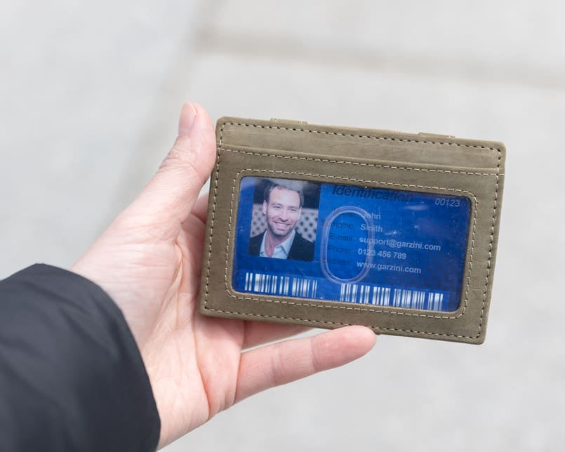 A hand showcasing the Olive Green Essenziale Magic Wallet with an ID Window. The hand holds a card inserted into the ID window of the wallet.