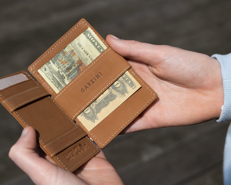 "Open Cavare Magic Wallet Vintage in Camel Brown with pull tab, and money straps with a hand holding the wallet open. "