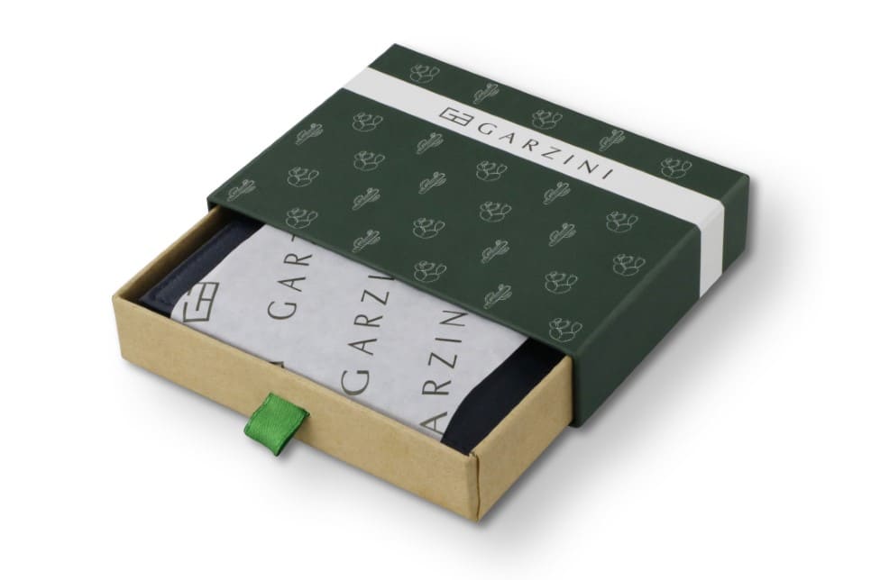 Half-open green box with Garzini brand name, featuring cactus icons. Inside the box, the Vegan Cactus Blue wallet is wrapped in tissue paper, placed in a light cardboard box with a green strap.