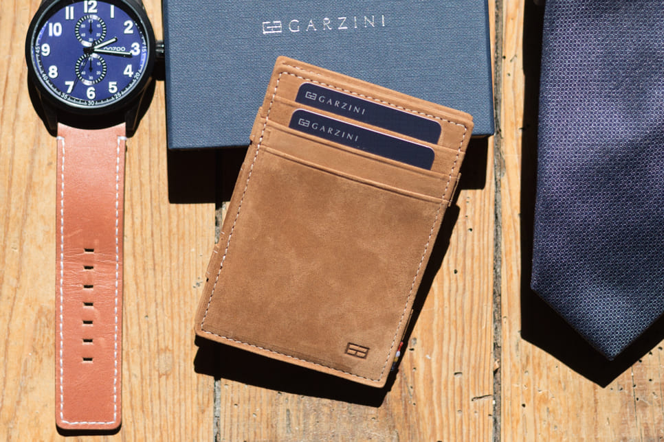 Front view of the Essenziale Magic Wallet Vintage in Camel Brown with three front card slots and two cards displaying the Garzini brand name. The wallet is placed on a Garzini packaging box, with a watch on the left and a tie on the right, both positioned on a wooden table