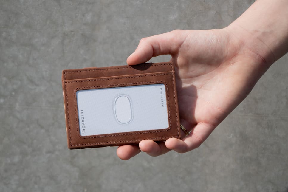 A hand showcasing the Brushed Brown Essenziale Magic Wallet with an ID Window. The hand holds a card inserted into the ID window of the wallet.