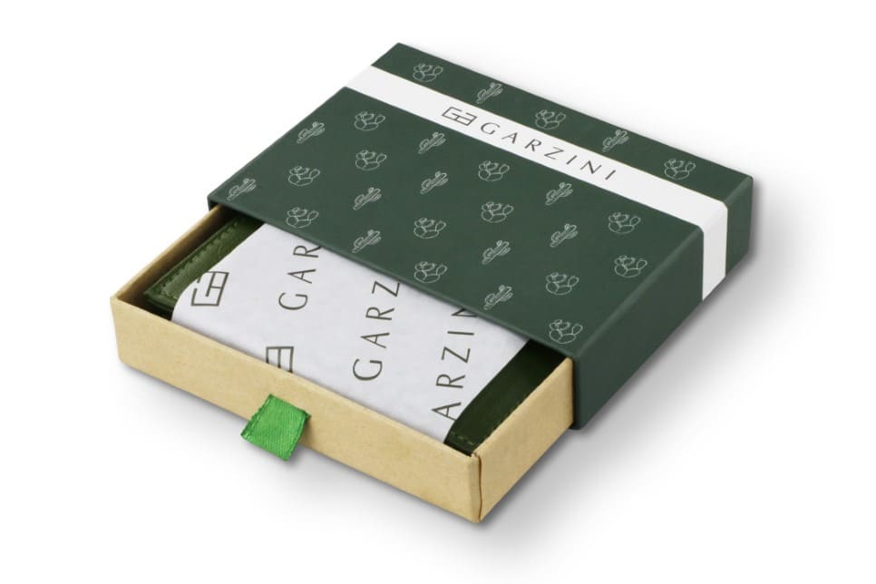 Half-open green box with Garzini brand name, featuring cactus icons. Inside the box, the cactus Green wallet is wrapped in tissue paper, placed in a light cardboard box with a green strap.