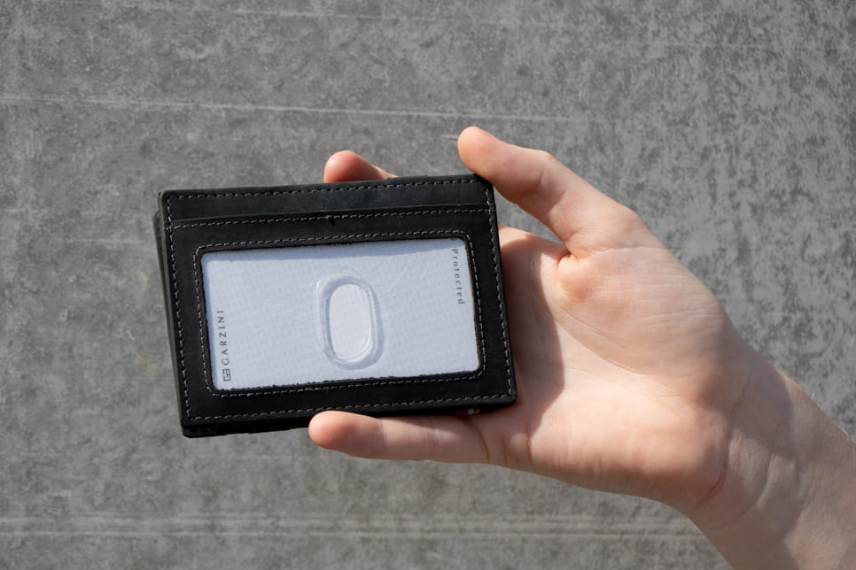 A hand showcasing the Carbon Black Essenziale Magic Wallet with an ID Window. The hand holds a card inserted into the ID window of the wallet.