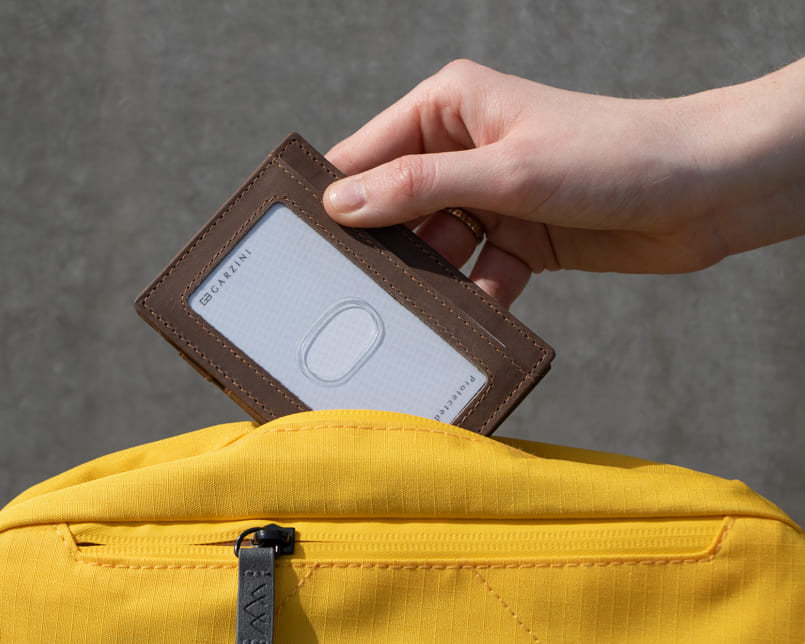 A hand grasping a Java Brown Essenziale Magic Wallet with an ID Window. The hand is inserting a card into the ID slot and placing the wallet inside a bag