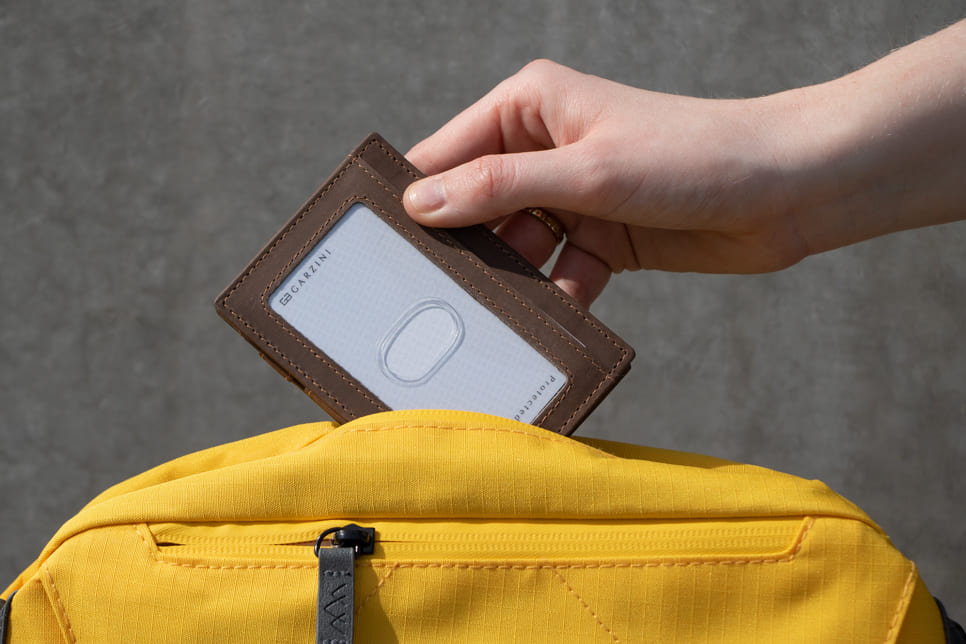 A hand grasping a Java Brown Essenziale Magic Wallet with an ID Window. The hand is inserting a card into the ID slot and placing the wallet inside a bag