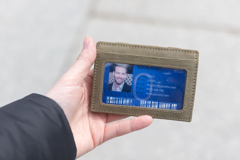 A hand showcasing the Olive Green Essenziale Magic Wallet with an ID Window. The hand holds a card inserted into the ID window of the wallet.