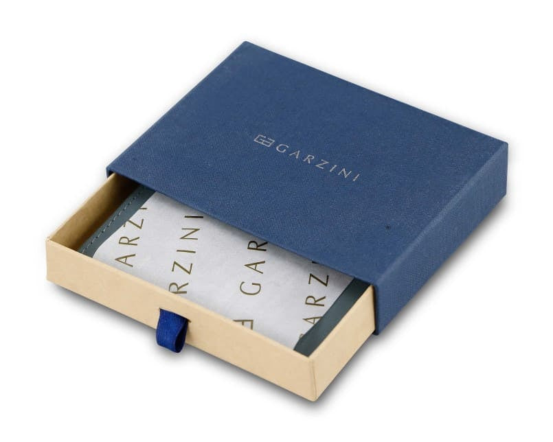 Half-open blue box with Garzini brand name. Inside the box, the Sapphire Blue wallet is wrapped in tissue paper, placed in a light cardboard box with a blue strap