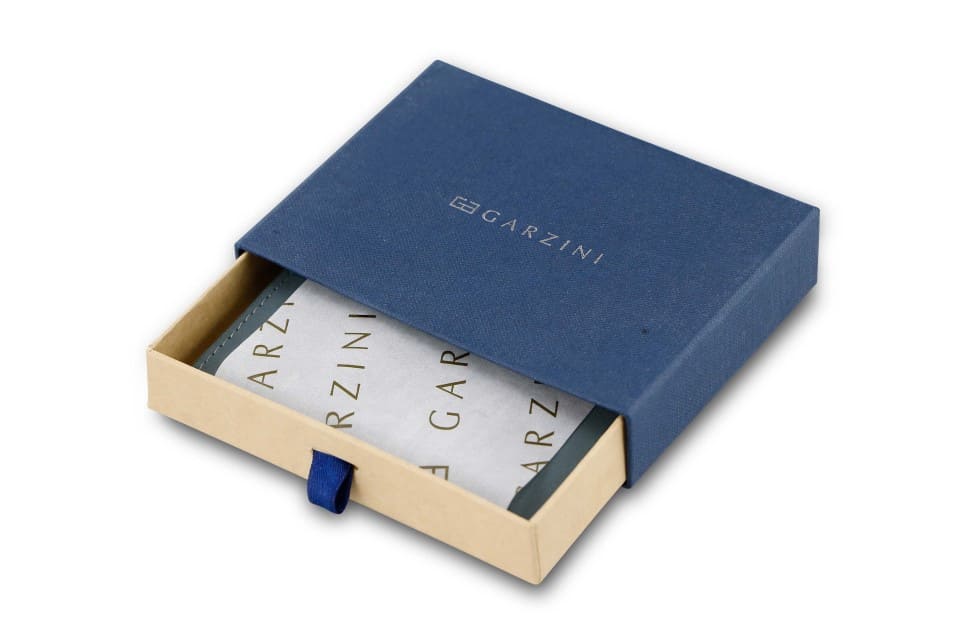 Half-open blue box with Garzini brand name. Inside the box, the Sapphire Blue wallet is wrapped in tissue paper, placed in a light cardboard box with a blue strap