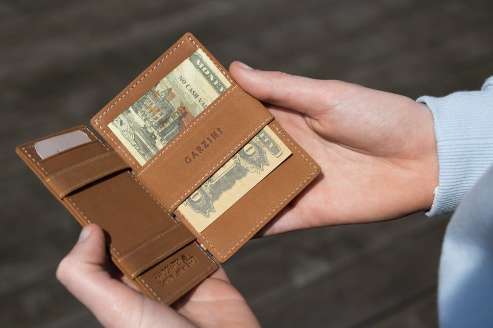 "Open Cavare Magic Wallet Vintage in Camel Brown with pull tab, and money straps with a hand holding the wallet open. "
